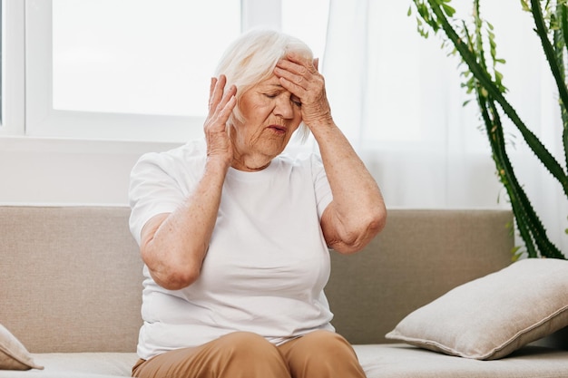 elderly woman severe pain in the head sitting on the couch health problems in old age poor quality of life grandmother with gray hair holding her head migraine and high blood pressure