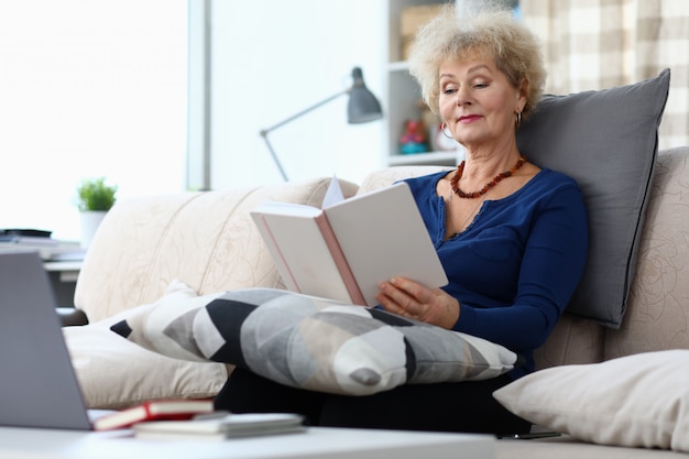 Elderly woman sits on sofa at home and reads book