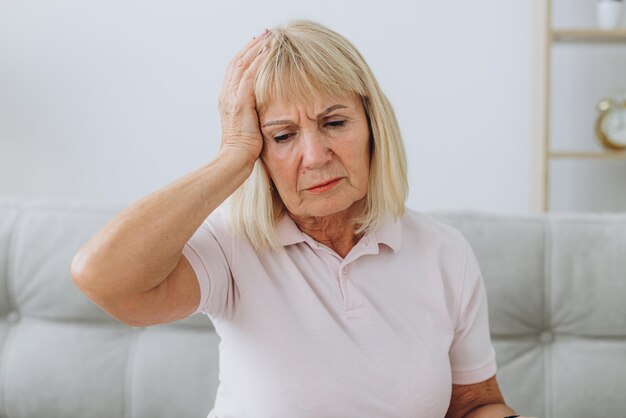 Фото Пожилая женщина сидит с головной болью и касается головы руками дома.