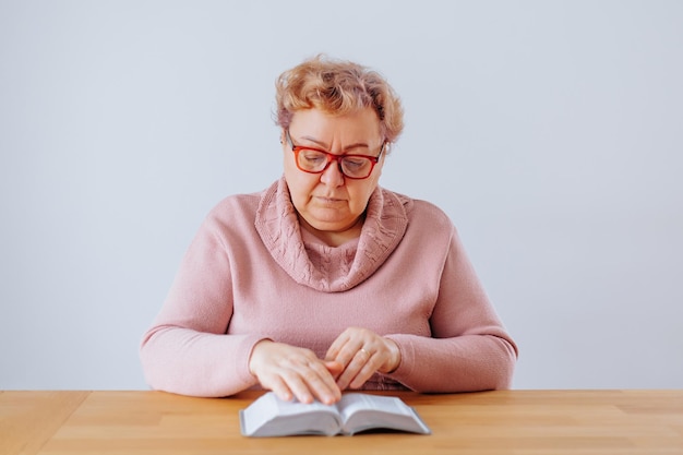 An elderly woman sitting in her home deeply engrossed in her book while wearing her glasses and enjo