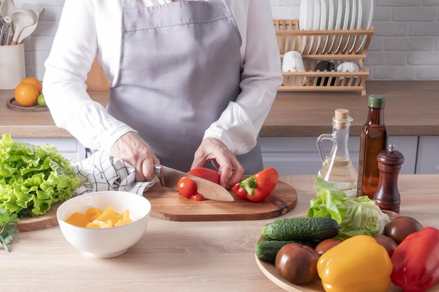 An elderly woman slices red fresh peppers on a cutting board closeup of the kitchen and countertops with vegetables in a bowl