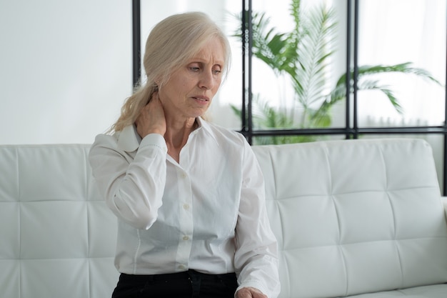 Пожилая женщина, страдающая от боли в шее, сидит на диване у себя дома
