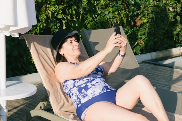 Elderly woman in swimsuit on sun lounger by the pool takes selfie on a smartphone