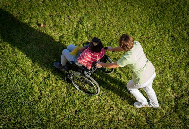 Elderly woman in wheelchair top view on grass with nurse taking care of her