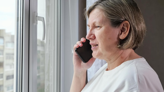 Elderly woman in white tshirt talks on phone near window