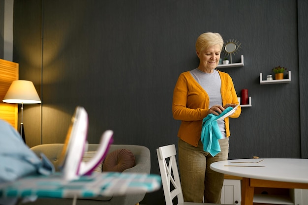 Elderly woman wiping dishware with cotton towel. Home living-room interior, domestic chore routine