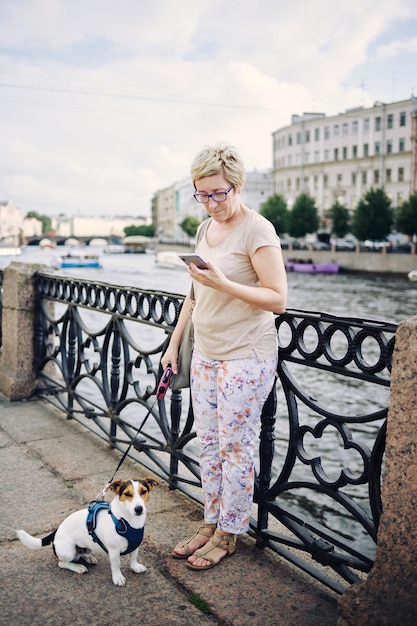 Elderly woman with a dog on a leash and looking at her phone near the river on a city street