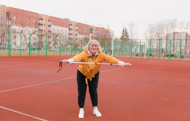 노란색 재킷을 입은 노인 여성이 빨간 디딜 방아에서 스포츠 연습을하고 있습니다. 경기장은 건강한 라이프 스타일입니다. 은퇴 한 사람과 스포츠. 활동적인 늙은 여자