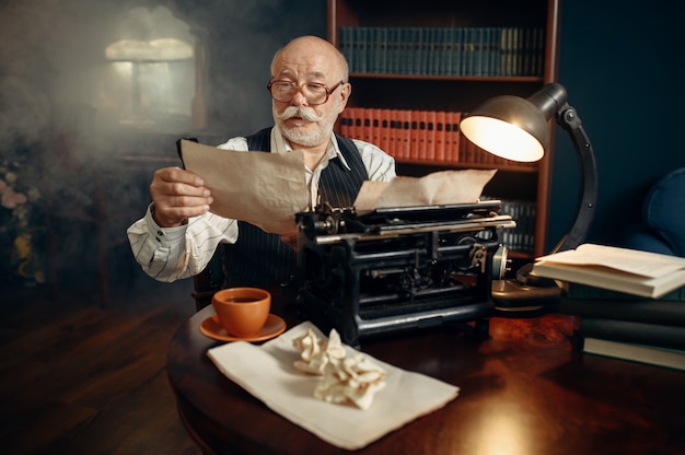 Elderly writer works on vintage typewriter in his home office. Old man in glasses writes literature novel in room with smoke