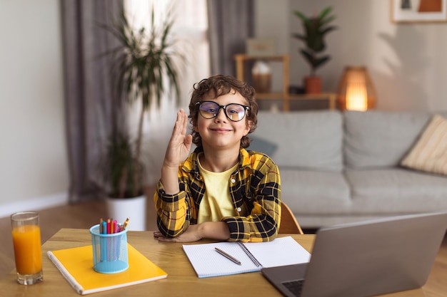 Elearning Cute schoolboy in eyeglasses raising hand and smiling to camera studying distantly at home free space