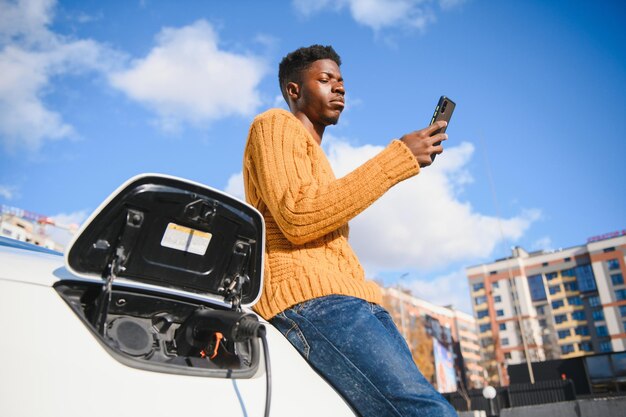 Electric cars, EV concept, eco friendly fuel. Portrait of young smiling black man, recharging his modern luxury electric car