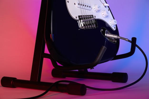 An electric guitar on a stand connected with a cord to an\
amplifier is illuminated with blue and red light. close-up.