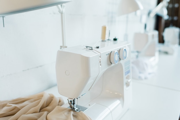 Electric white sewing machine with piece of beige fabric on desk inside workshop of contemporary seamstress or fashion designer