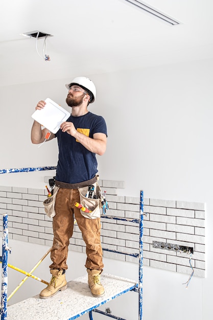 Electrician Builder with beard worker in a white helmet at work, installation of lamps at height.