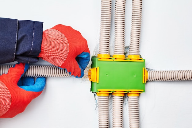 An electrician connects an electric conduit to square-shaped distribution box made of green plastic and containing eight ways.