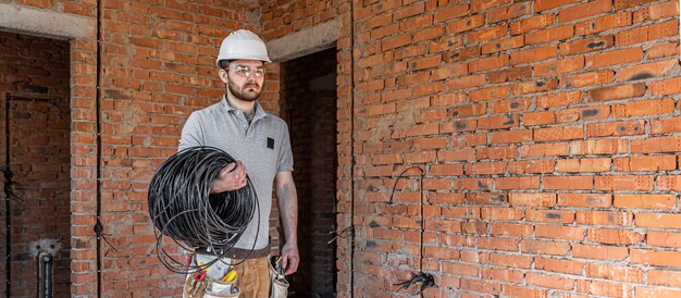 An electrician in a hard hat looks at the wall while holding an electric cable.