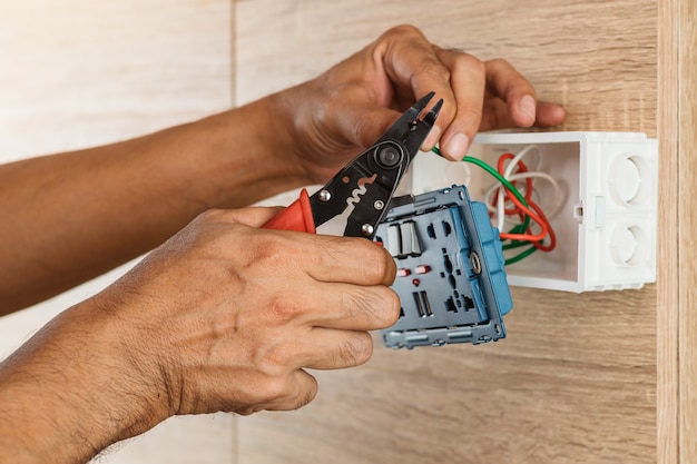 Photo electrician is stripping electrical wires in a plastic box on a wooden wall to install the electrical outlet.