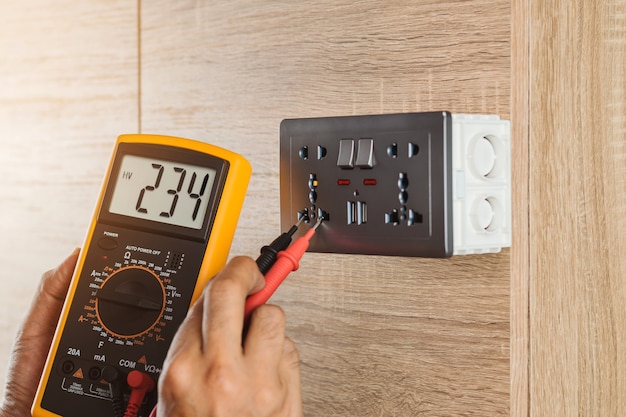 Photo electrician using a digital meter to measure the voltage at a wall socket on a wooden wall.