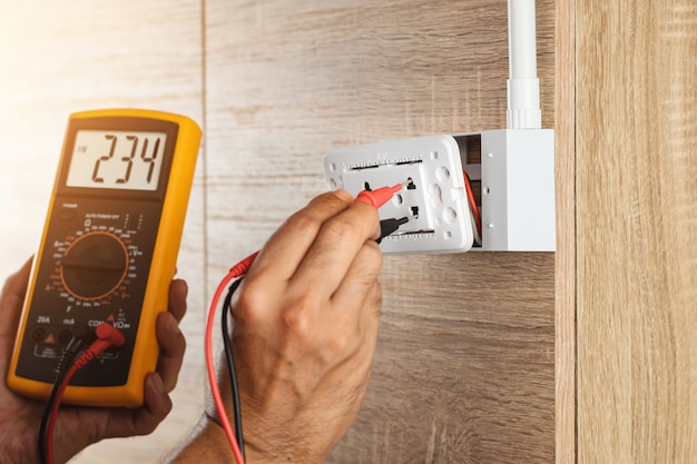 Photo electrician using a digital meter to measure the voltage at a wall socket on a wooden wall.