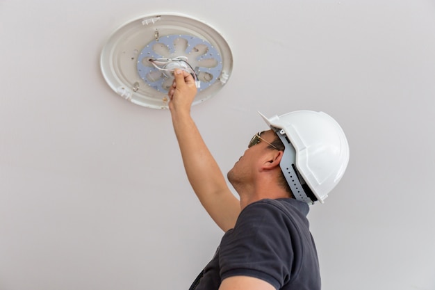 Elettricista con il casco bianco che controlla illuminazione al soffitto nella casa, concetto del tecnico.