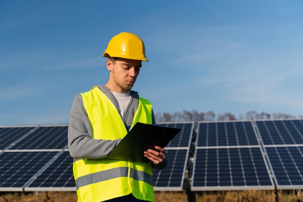 Electricity engineer man holding clip board and taking notes\
while looking at solar panels green energy and ecology concept