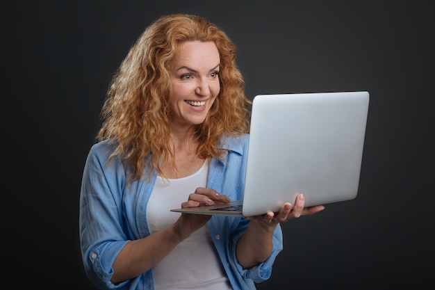 전자 통신. 회색 배경에 고립 된 서있는 동안 그녀의 친구와 의사 소통을 위해 그녀의 노트북을 사용하는 영감을받은 귀여운 동기 부여 여성
