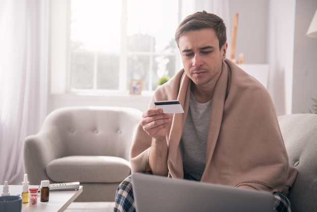 Electronic payment. Sad ill man looking at the laptop screen while holding a credit card in his hand