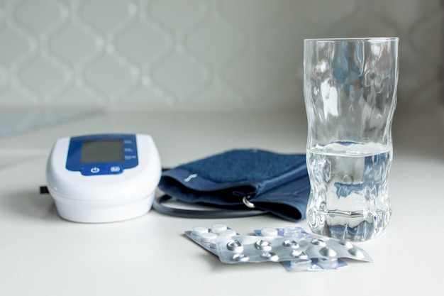 Electronic tonometer, pills and a glass of water on a white table