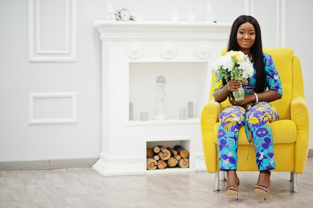 Elegance african american woman model wear in colored clothes Chic afro women in room sitting on yellow chair with flowers in hand