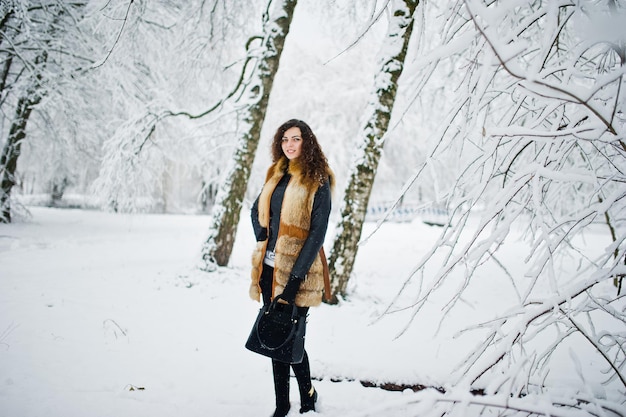 겨울에 눈 덮인 숲 공원에서 모피 코트와 핸드백에 우아한 곱슬 소녀.