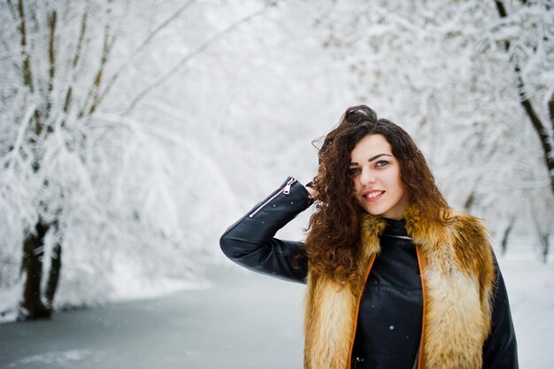 Elegance curly girl in fur coat at snowy forest park at winter.