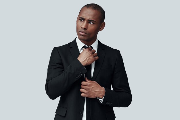 Elegance. Handsome young African man in formalwear adjusting necktie while standing against grey background