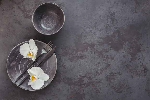 Elegance table setting with knitted grey napkin, cutlery, ceramic plates, glasses and white orchid flowers on dark table. Holiday modern table decoration. Romantic dinner.