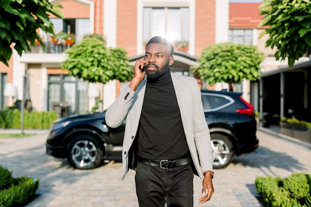 電話、黒い車のクロスオーバー、建物を取っている間通りを歩いてエレガントなアフリカの実業家