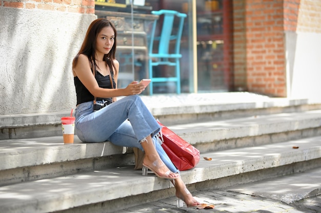 Elegant asian woman sitting on stairs in front of shop holding mobile phone posing lifestyles