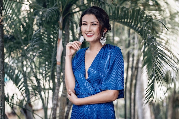 Elegant Asian woman wearing blue low-neck dress shows her massive earrings; commerce concept.