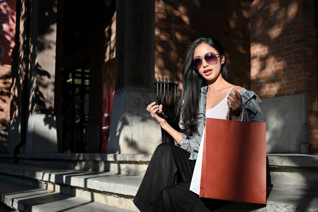 Элегантная азиатка в солнцезащитных очках с сумками для покупок сидит на уличной лестнице