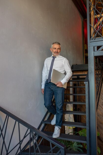 Elegant attractive gray-haired man with the computer walking downstairs