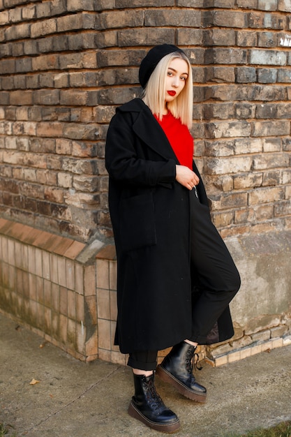 革のブーツの赤いシャツの黒いベレー帽のスタイリッシュなロングコートでエレガントな魅力的な若い女性のブロンドは、街のヴィンテージのレンガの壁の近くに立っています