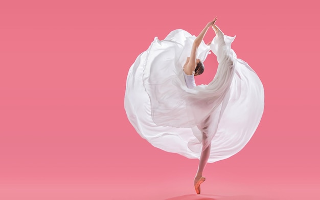 Elegant ballerina in pointe shoes dancing in a long white skirt on a pink background