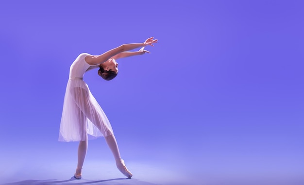 An elegant ballerina in pointe shoes is dancing in a long flying white skirt on a lilac light background