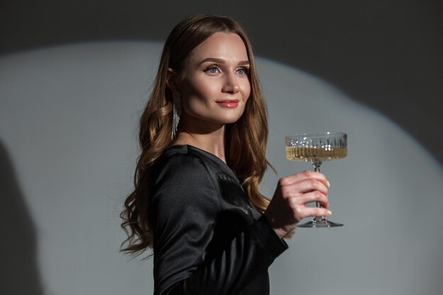 Elegant beautiful chic lady with an earring in a trendy fashion black dress with a vintage champagne glass celebrating an event on a dark background with circular light Pretty woman