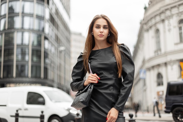 Elegant beautiful fashionable business woman in black stylish clothes with a trendy dress and a fashionable leather black handbag walks in a modern city