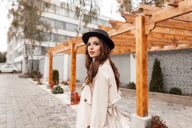 Elegant beautiful young curly girl with a stylish hat in a fashionable beige classic coat walks on the street on an autumn day