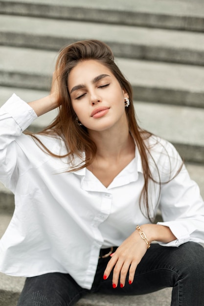 Elegant beautiful young woman model in fashion casual clothes with a white shirt and black jeans stands near a vintage building in the city Pretty girl