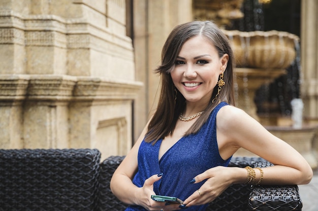 Elegant beautiful young woman with a smartphone in her hands looks at the camera while sitting in a street cafe