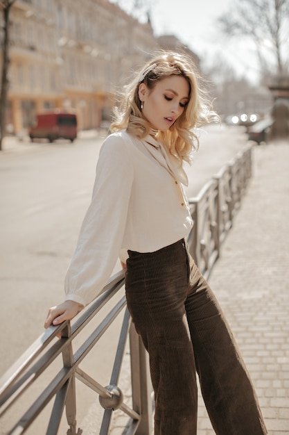 Elegant blonde curly woman in stylish velvet brown pants and trendy white blouse looks down and leans on fence on city street
