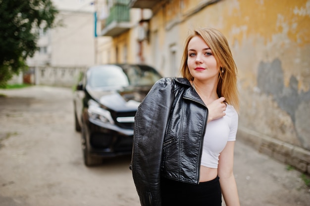 Elegant blonde girl wear on black leather jacket posing at streets of town, luxury car.