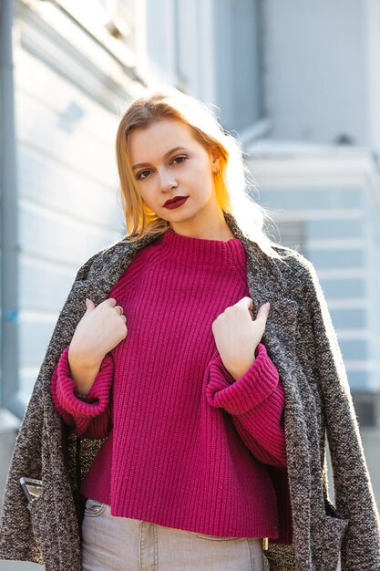 Elegant blonde woman wearing coat and sweater, posing with soft backlight at the city