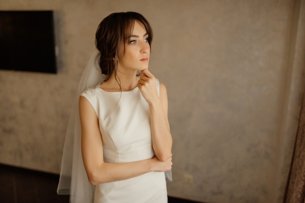 Elegant bride stands near the window in a wedding dress at hotel. Wedding morning of the bride.